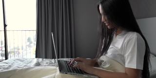 一个年轻的女人坐在房间的床上，用他们的笔记本电脑工作。工作，网上购物，查看电子邮件或在家里的床上搜索信息。