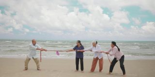 夏天，亚洲的老男人和老女人在海滩上和朋友玩拔河比赛。到了退休年龄去海边度假。友谊和团队合作理念。慢动作镜头
