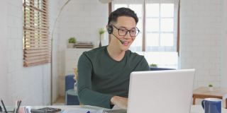 微笑英俊的亚洲商人戴着耳机在家远程工作。他是一个网络视频会议