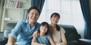幸福的亚洲家庭呆在客厅里一起看电视电影。父亲和母亲花闲暇时间与小女孩女儿坐在沙发上，看动画片，在家里一起欢笑。