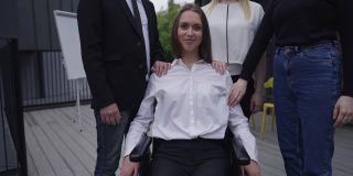 正面的观点肖像快乐的残疾年轻女子在轮椅看镜头微笑触摸的手的同事在肩膀上。白人截瘫员工在户外与同事摆姿势。