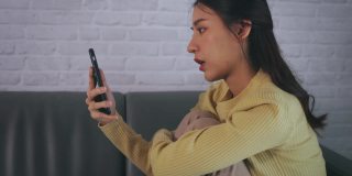 年轻而疲惫的亚洲女性焦虑不安地坐在家里的沙发上，一边看手机一边等待电话或新闻。