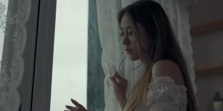 4K沮丧美丽的亚洲女人独自站在公寓房间的窗口哭泣和尖叫。孤独悲伤的女孩，想起悲伤烦恼。消极情绪与心理健康的概念。