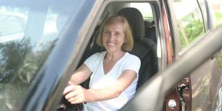 快乐的老妇人驾驶着坐在崭新的棕色轿车里，微笑着看着相机享受旅程。驾驶课程和人寿保险概念。退休人员活动理念。