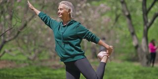 快乐的资深女运动员站在户外单腿伸展肌肉。热身运动的积极自信适合高加索退休人员在阳光明媚的春天夏天公园。生活方式和运动概念