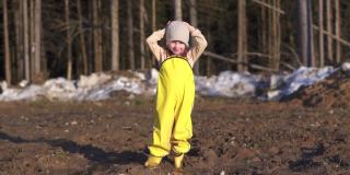 一个小女孩在春天的村子里跳泥巴。童年快乐女儿开心。森林里覆盖着融化的积雪。连身衣可以保护脏衣服。在阳光明媚的日子里，穿着黄色的胶靴在地上跳舞