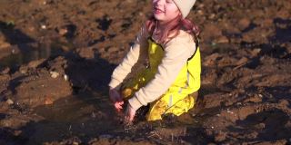 可爱的孩子坐在泥泞的水坑里玩手。小女孩在春天的村庄泥。快乐的童年的女儿。森林里潮湿的地面上有融化的雪。黄色的连身衣和橡胶靴保护着你。