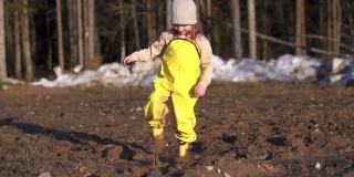 女孩走到泥里。在地上的水坑里跺脚玩游戏。快乐的孩子在乡村森林里玩春天。黄色防水连身衣，胶靴。溅起的尘土飞向两侧