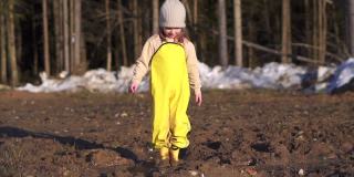 女孩正在往泥里跳。地上的水坑用来玩游戏。在森林附近的村庄里，一个快乐的孩子在春天里玩耍。黄色防水连身衣和胶靴。