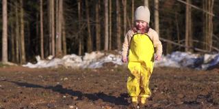 女孩正在往泥里跳。地上的水坑用来玩游戏。在森林附近的村庄里，一个快乐的孩子在春天里玩耍。黄色防水连身衣和胶靴。