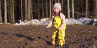 女孩的乐趣是跳进泥里。地上的水坑用来玩游戏。在森林附近的村庄里，一个快乐的孩子在春天里玩耍。黄色防水连身衣和胶靴。