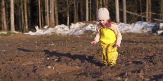 可爱的孩子坐在泥泞的水坑里玩手。小女孩在春天的村庄泥。快乐的童年的女儿。森林里潮湿的地面上有融化的雪。黄色的连身衣和橡胶靴保护着你。