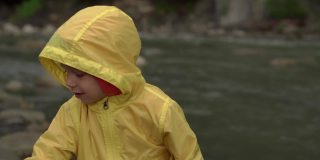 山区河流附近的学龄前儿童。穿黄色雨衣的小孩向河里扔石头。男孩把鹅卵石从岸上扔进水里。度假，去露营，春天，野性的自然，家庭观念