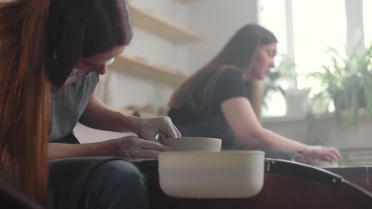 两个年轻的女人正在工作与陶轮制作锅在大师班为成年人，创造性的工匠爱好。DIY休闲活动和工艺概念。慢动作