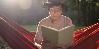阳光明媚的日子里，一个戴着眼镜和帽子的年轻人坐在吊床上看书