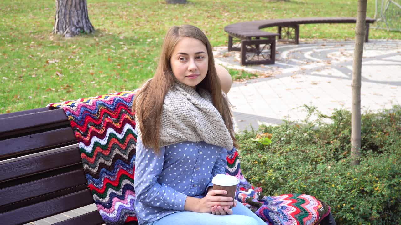 美丽的女孩坐在长椅上，在一个阳光明媚的秋天公园。女孩在户外喝着热咖啡。秋天日落时，年轻的黑发女子坐在长凳上
