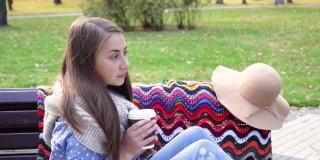 美丽的女孩坐在长椅上，在一个阳光明媚的秋天公园。女孩在户外喝着热咖啡。秋天日落时，年轻的黑发女子坐在长凳上