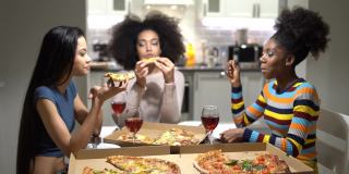快乐的三名多民族的年轻女子坐在厨房里，吃着披萨，喝着葡萄酒，讲着笑话，放声大笑