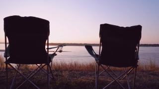 一对游客椅，一个折叠椅，在河边的背景下，美丽的夕阳，放松和休息，享受与自然的统一。视频素材模板下载