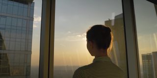 透过摩天大楼的窗户看日落的女人的背影