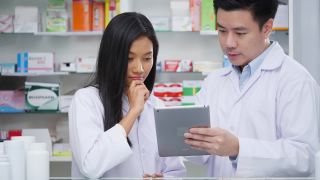 图为在医院药房检查药品的男女药剂师的照片。亚洲医生正在检查药片上的药物数据，穿着医疗外套。有药店药学背景的货架。视频素材模板下载