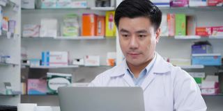 男药师在医院药房网上会议。亚洲男人医生建议使用药物和笔记本电脑。有药店药学背景的货架。