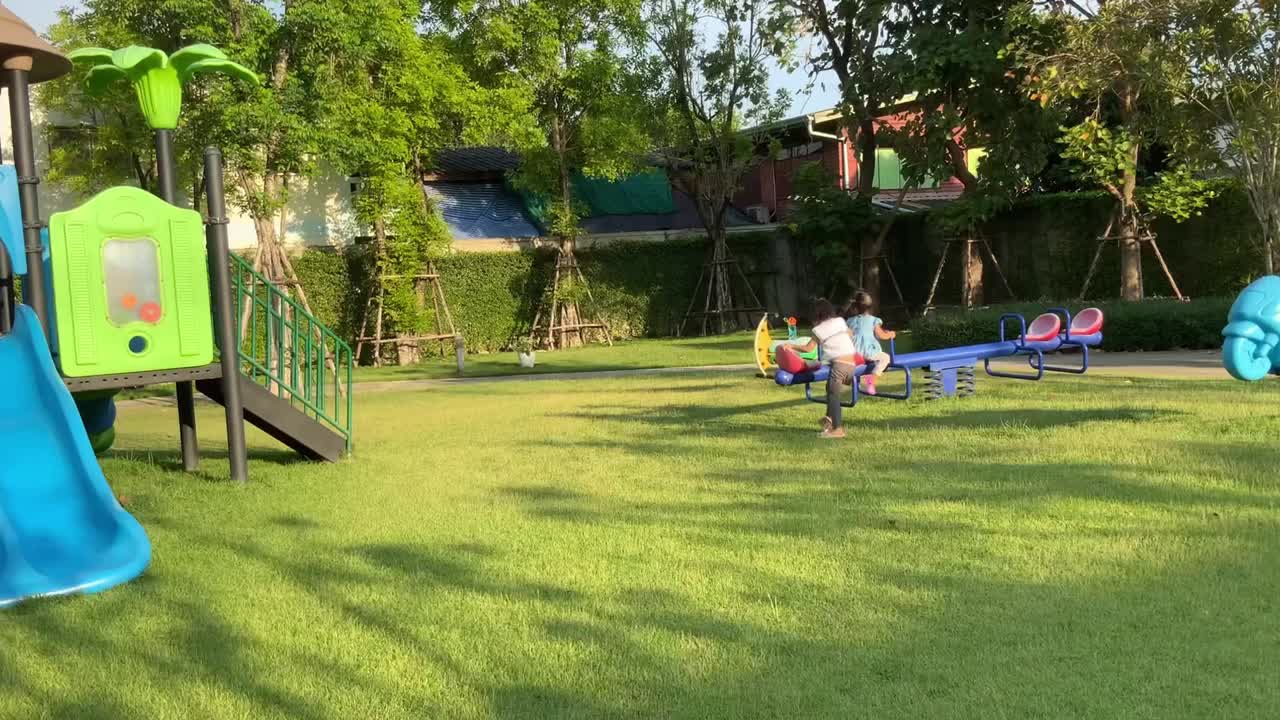 两个亚洲小女孩泰国兄弟姐妹一起在一个大的花园操场上玩跷跷板。孩子们在许多大树和绿草下进行户外运动。