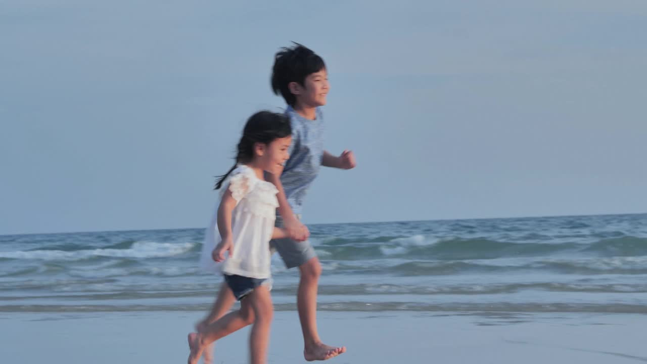 幸福两个4-8岁的亚洲男孩和女孩手牵着手，在海边的沙滩上快乐地奔跑。活跃的孩子户外活动在热带暑假。援助之手的概念。