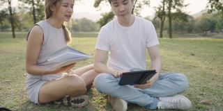 年轻的大学同学们坐在公园里的地上，用数码平板电脑讨论小组作业，朋友们一起脑力激荡工作，轻松的会议环境温暖阳光