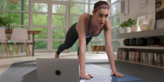 女运动员用笔记本电脑在瑜伽垫上做平板支撑。在家锻炼