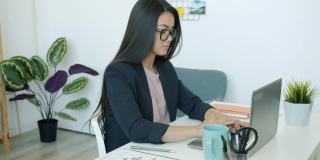 亚洲女员工穿着西装，在办公室用笔记本电脑工作，喝咖啡，看文件