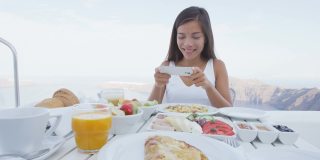 一个女人用手机app拍早餐。一个女孩用社交媒体拍豪华旅行度假的食物。欧洲希腊圣托里尼度假胜地的美女。