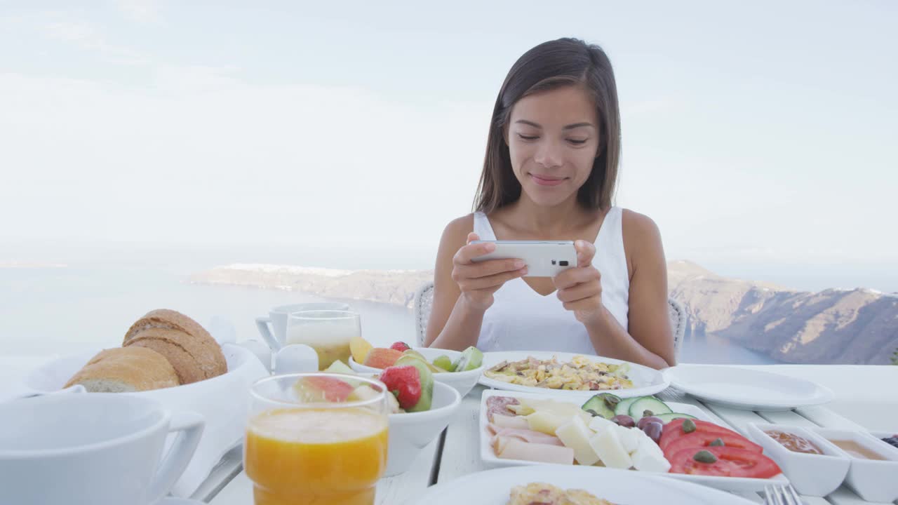 美女用手机app吃早餐。餐桌上有各种各样的健康食品。女游客正在圣托里尼岛的露台度假胜地放松。