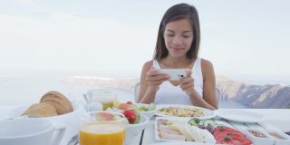 美女用手机app吃早餐。餐桌上有各种各样的健康食品。女游客正在圣托里尼岛的露台度假胜地放松。