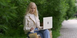 一个女学生带着笔记本电脑和书坐在公园的长椅上。一名年轻女子在绿树的背景下翻阅笔记本电脑上的书籍和印刷品。