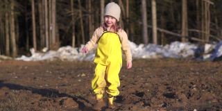 快乐的孩子在泥泞的水坑里跳舞。村子里春天的小女孩站在泥里。快乐的童年跳舞的女儿。潮湿的地面融化了森林里的雪。连身衣可以保护脏衣服。