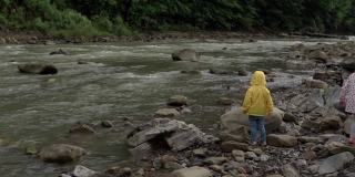 小学前班的孩子在山河附近。穿黄色雨衣的小孩向河里扔石头。男孩从岸上把鹅卵石扔进水里。度假，去露营，春天，野生自然，家庭观念