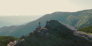 一个徒步旅行者站在一块岩石上，在日落时展望未来。运动员站在山顶眺望远方。从后面鸟瞰一个圆圈