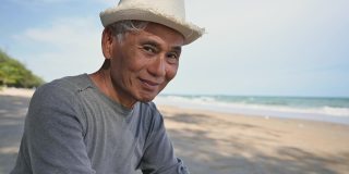 亚洲老人戴着帽子坐在沙滩上微笑着用手势打电话聊天。