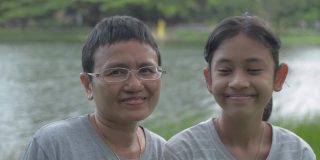 快乐的亚洲单身妈妈戴着眼镜，和她可爱的女儿站在公园的自然池塘边看着相机，摆出亲密的姿势。