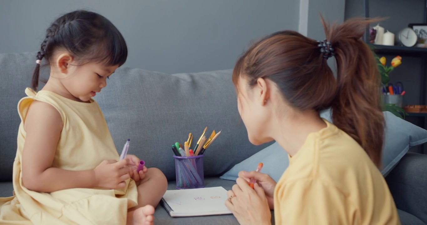 快乐快乐的亚洲家庭妈妈教女孩画画，使用相册和彩色铅笔在家里客厅的沙发上放松。