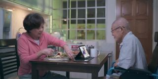 一位坐着轮椅、戴着气管造口管的亚洲老人，在晚餐后的闲暇时间，妻子在厨房递给他苹果时，他用数码平板电脑给家人打电话