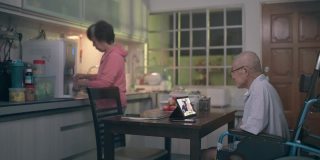 坐在轮椅上的亚洲老人和妻子在晚餐后的闲暇时间在厨房用平板电脑给家人视频通话