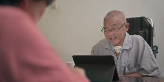 一位坐着轮椅、戴着气管造口管的亚洲老人，在晚餐后的闲暇时间，妻子在厨房递给他苹果时，他用数码平板电脑给家人打电话
