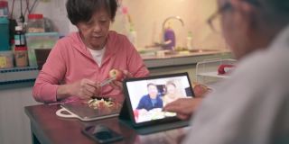 一位坐在轮椅上的亚洲老人在晚饭后的闲暇时间用数码平板电脑给家人打视频电话，而妻子则在厨房递给他苹果