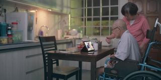 坐在轮椅上的亚洲老人和妻子在晚餐后的闲暇时间在厨房用平板电脑给家人视频通话