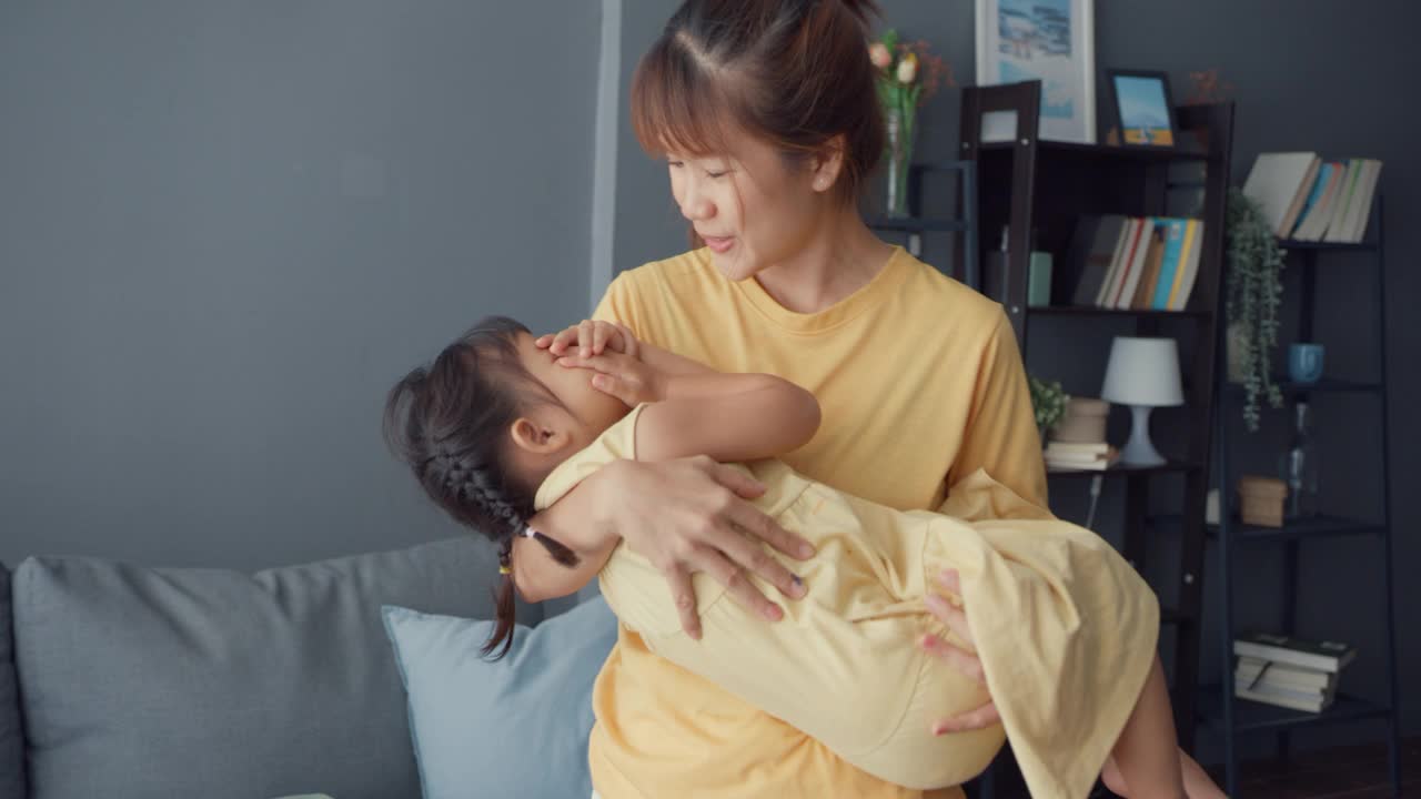 快乐开朗的亚洲家庭妈妈抱着蹒跚学步的小女孩在怀里哭泣，在家里的客厅里安慰呵护着。
