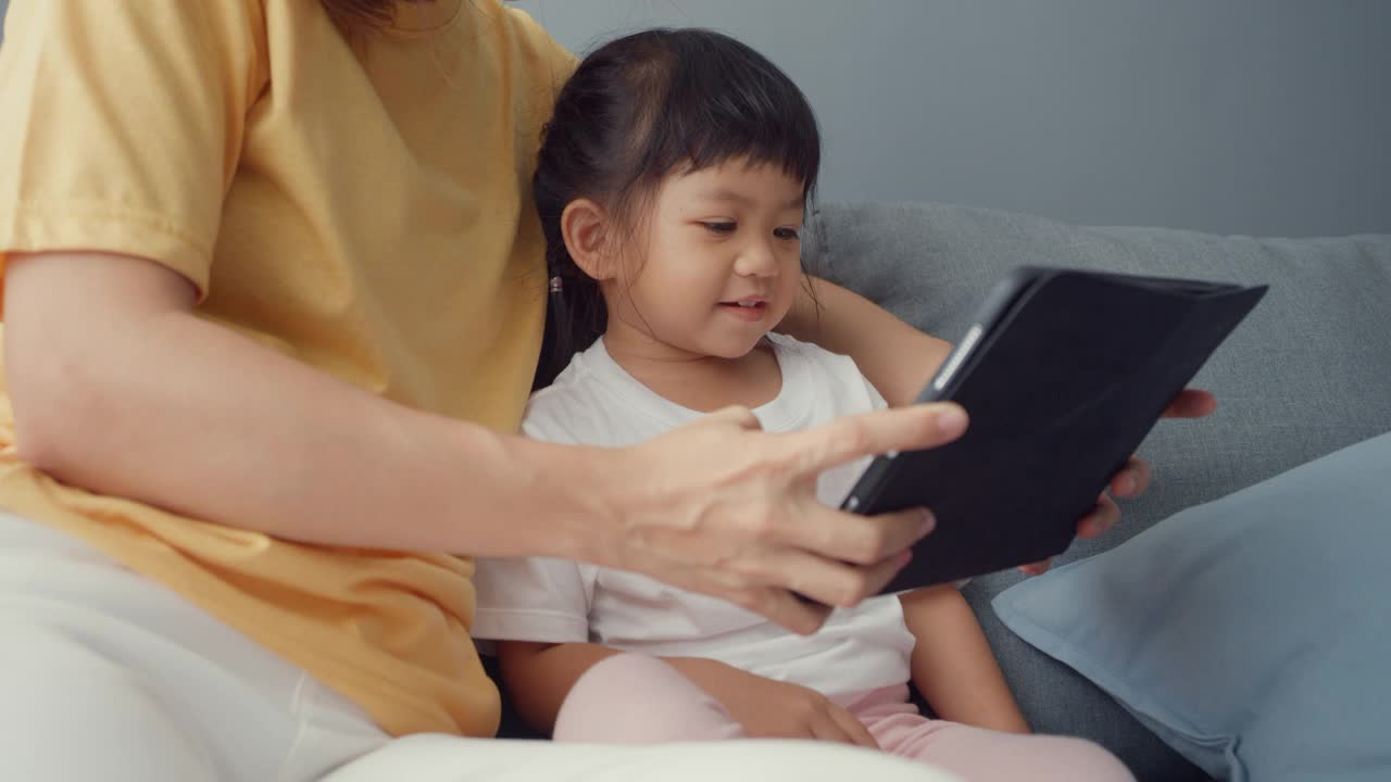 快乐快乐的亚洲家庭妈妈和可爱的孩子使用数字平板电脑兴趣卡通和看电影，在家里客厅的沙发上放松。