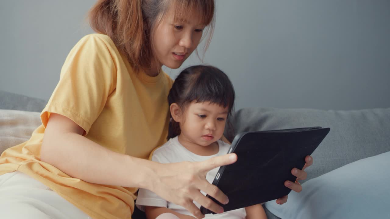 快乐快乐的亚洲家庭妈妈和可爱的孩子使用数字平板电脑兴趣卡通和看电影，在家里客厅的沙发上放松。