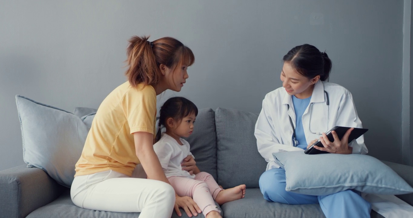年轻的亚洲女儿科医生和小女孩病人使用数字平板电脑分享良好的健康测试消息与快乐的妈妈坐在沙发在家里。